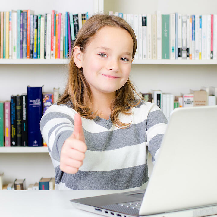 Ein junges Mädchen sitzt an einem Laptop vor einem Bücherregal und streckt den Daumen in die Kamera.